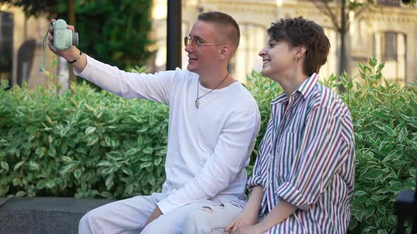 Side View Cheerful Young Couple Taking Selfie with Retro Camera Sitting on City Street Outdoors alt