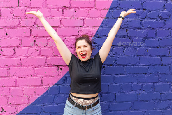 Cheerful positive young woman in goth clothing posing against a blue-purple brick wall. The concept