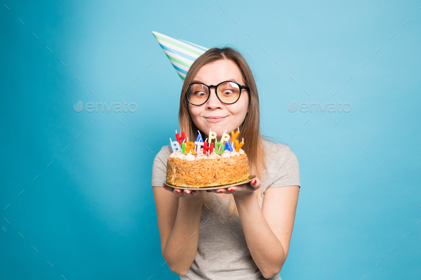 Crazy funny girl in a paper hat and glasses holding a big birthday cake on  the blue background Stock Photo by Satura_