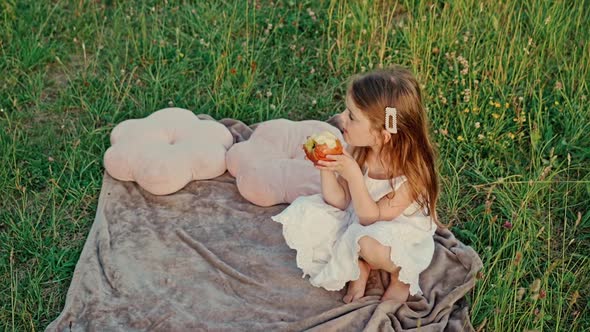 Little Cute Girl with Flowing Hair in a White Summer Dress Lying on a Green Lawn Eating an Apple