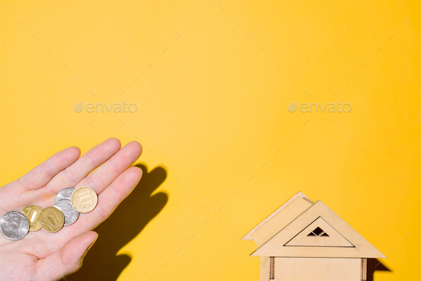 Toy house and man\'s hand holding coins, top view. Yellow background, copy space. Concept of rent