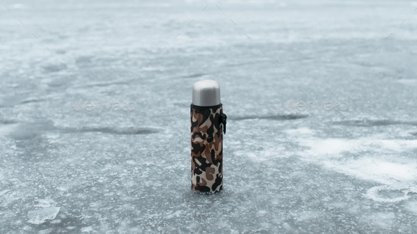 Thermos in a khaki case standing on a frozen lake. Concept hot drink in cold weather, winter fishing