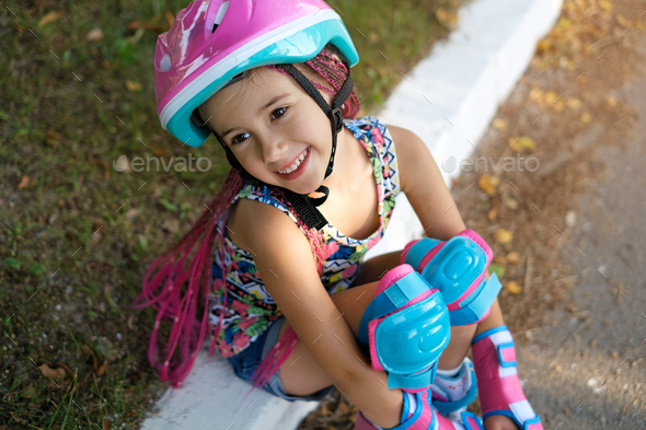 Girl in a helmet and knee pads sat side of the sidewalk after roller skating.