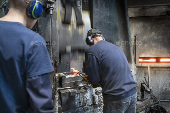 Engineer with apprentice forging steel parts in hammer press in industrial forge
