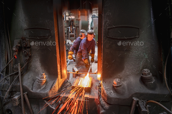 Engineer with apprentice forging steel parts in hammer press in industrial forge