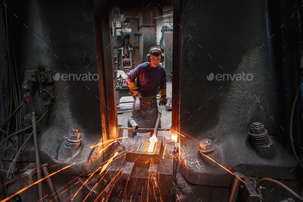 Apprentice engineer forging steel parts in hammer press in industrial forge