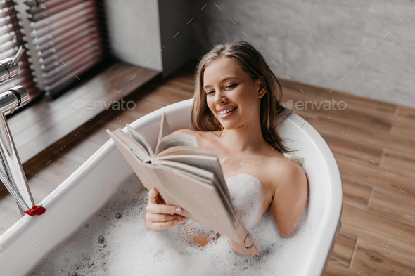 Young lady lying in foamy bath and reading book in relaxing atmosphere at home bathroom, free space