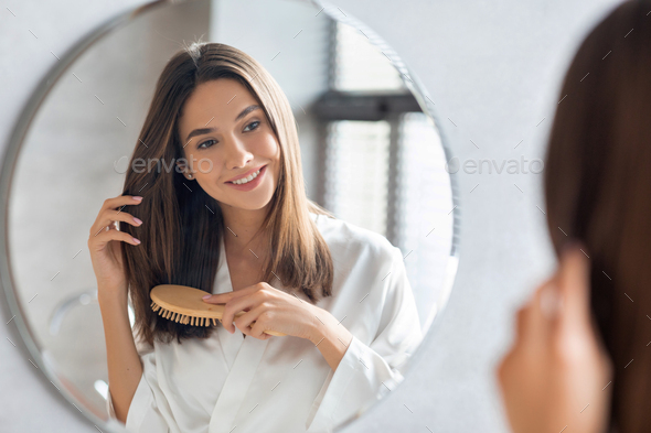 Haircare Concept. Attractive Young Lady Brushing Her Hair With Comb Near Mirror