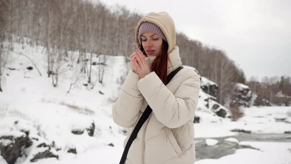 A Beautiful Girl in a Warm Jacket and a Hood is Trying to Warm Frozen Hands
