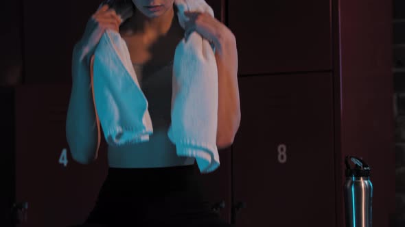 A Young Pretty Woman Sitting in Locker Room with a Towel on Her Neck  Looking in the Camera alt