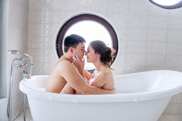 Close-up portrait of couple face to face in bubble bath at round window overlooking the sea.