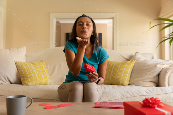 Portrait of african american woman holding valentine\'s gift blowing kiss on video call at home