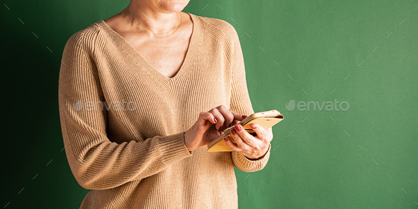 Cropped shot of woman in light jumper holding phone her hands against green wall