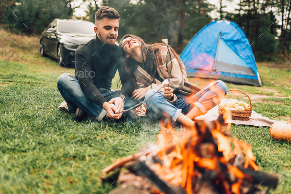 Young couple in nature near the campfire roasting marshmallows and laugh cheerfully
