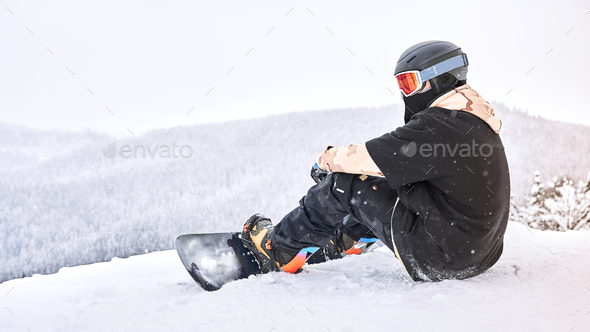 Male snowboarder sitting on the hilltop while snowing and take a rest for a while - Stock Photo - Images