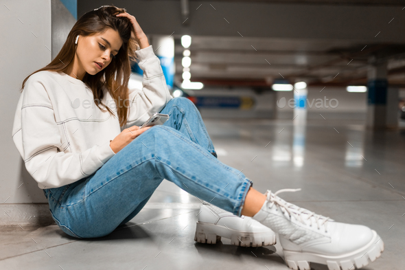 Stylish girl in the underground parking sits on the floor and listens to music