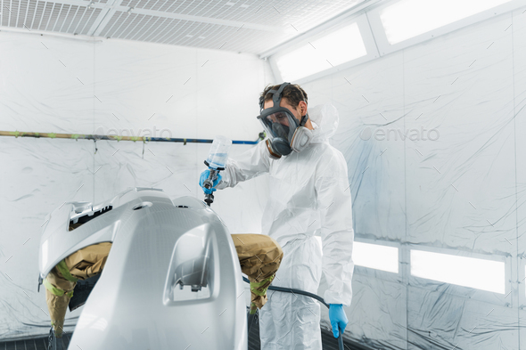 Professional car painter in a protective suit and mask varnishes a painted bumper of a vehicle