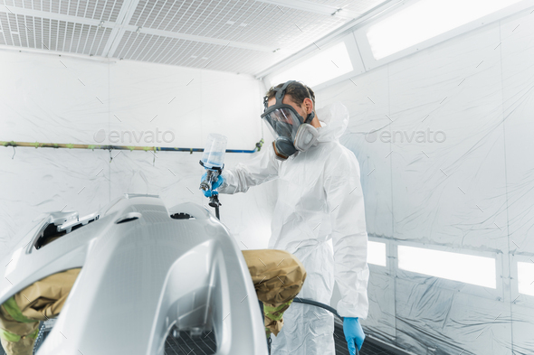 Professional car painter in a protective suit and mask varnishes a painted bumper