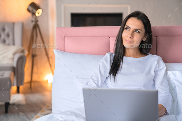Smiling beautiful woman using notebook while lying in bed at home with dreaming look away