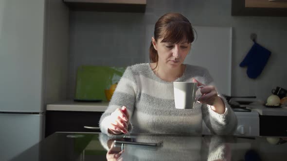 Pensive Woman Sitting in the Kitchen at Home with a Cup of Tea and Uses the Phone