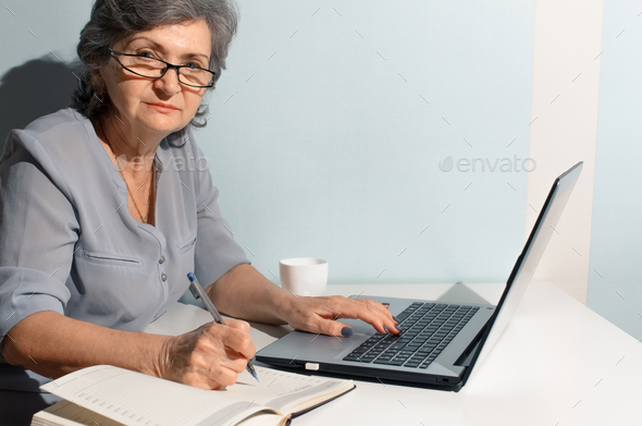 Elderly woman using laptop and writing in diary. Senior woman porter looking at camera