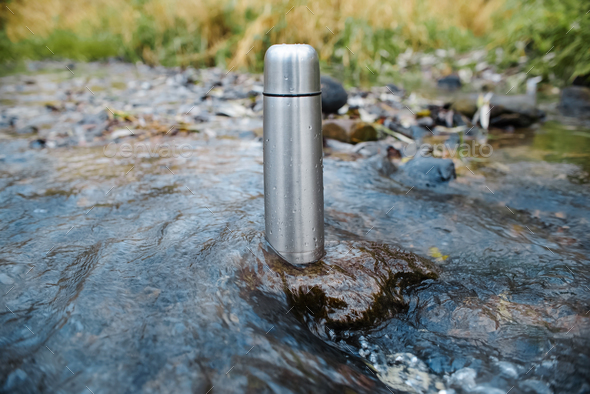 Traveling steel thermos stands in the middle of the stream. Mountain stream and vacuum flask.