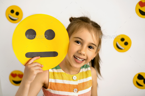 A happy, cheerful girl looks out from behind a unhappy cardboard smile face - Stock Photo - Images