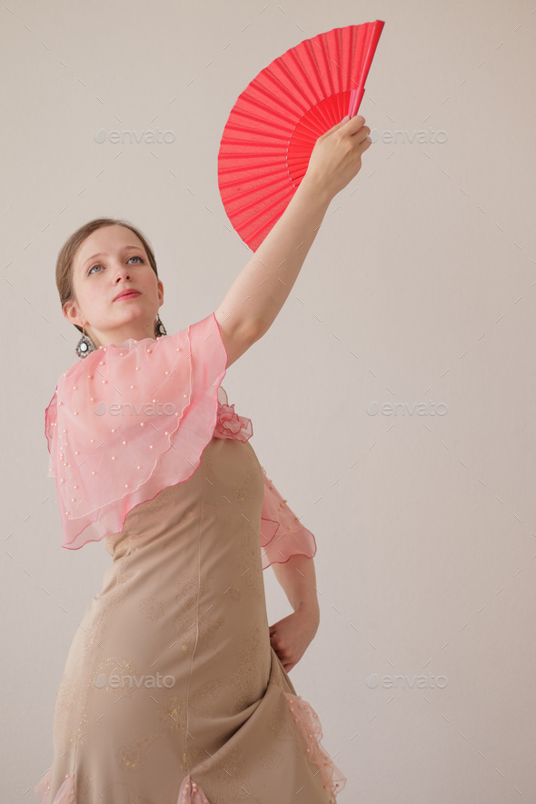 Portrait of beautiful young woman in beige, pink dress dancing flamenco.Hand with red fan raised up.