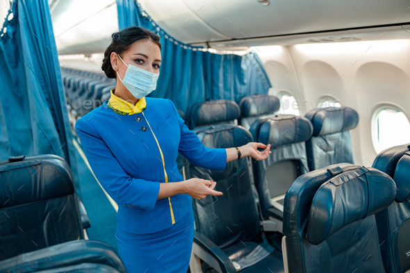 Stewardess in medical mask inviting to take passenger seat in plane