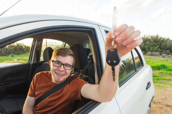 Portrait of happy smiling young man, buyer sitting in his new car and showing keys outside dealer