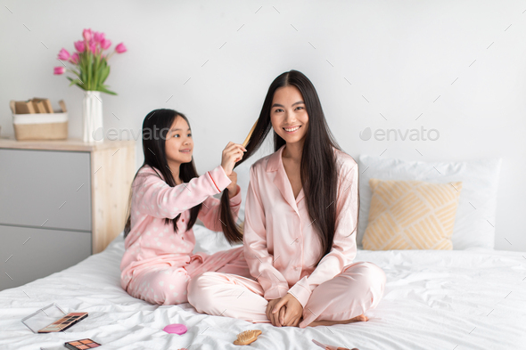 Cheerful korean teenage girl combing long smooth hair to millennial woman, sit on bed with cosmetics