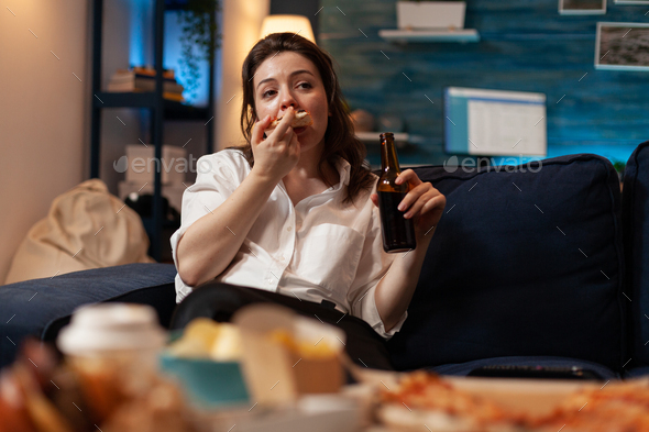 Woman eating a slice of hot pizza delivery sitting on couch holding beer bottle looking at