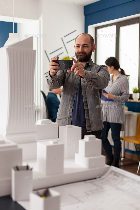 Smiling design architect holding smartphone to photograph desk with architectural model