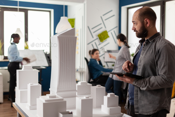 Architect holding tablet with blueprints inspecting design of white foam building model