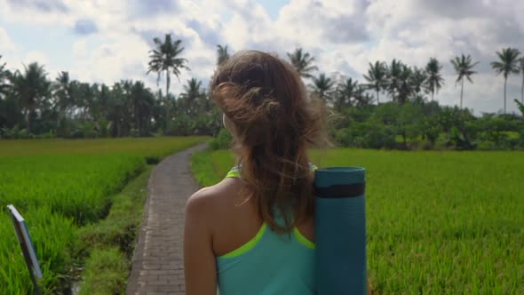 Slowmotion Steadicam Shot of a Young Woman with Yoga Math Walking Through a Beautiful Rice Field alt