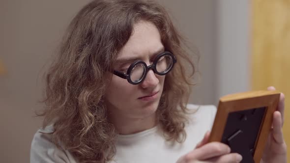 Sad Young Caucasian Man in Retro Eyeglasses with Long Curly Hair Looking at Photograph and Crying alt