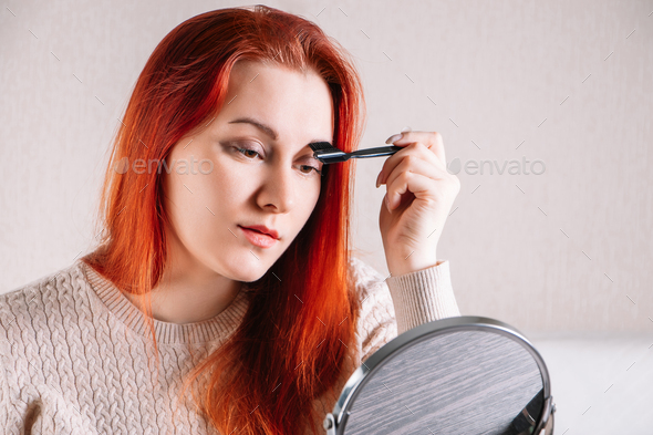 Young woman with red hair applies makeup. Lady looks in mirror and use cosmetics