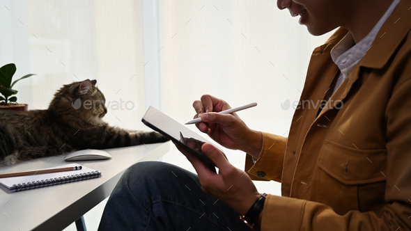 Cropped shot of casual man holding pen wiring on notebook and sitting with lovely cat at home.