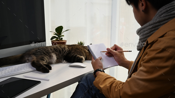 Young man holding pen wiring on notebook and sitting with cute cat at comfortable home.
