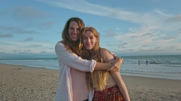 Mother and Teenage Daughter at Beach Hugging and Smiling at Camera alt