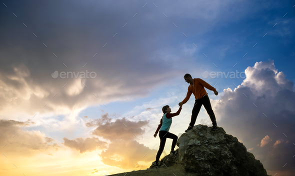 Man and woman hikers helping each other to climb a big stone at sunset in mountains. Couple climbing