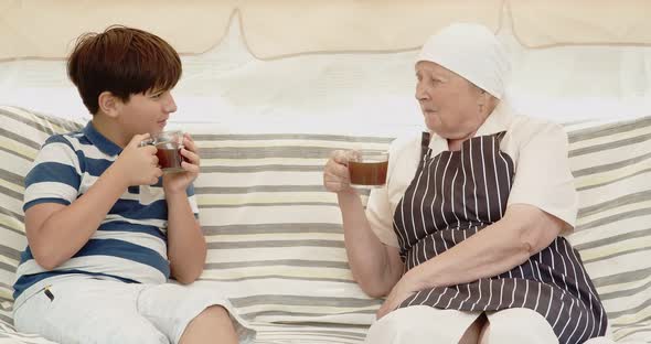 A Teenager and His Grandmother are Drinking Tea While Sitting on a Garden Swing alt