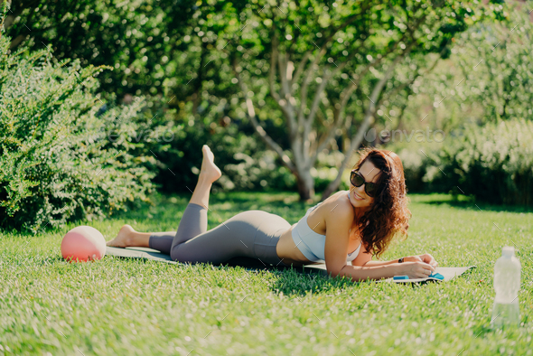 Sporty woman lying on karemat wears activewear raises leg has bare feet  poses on green grass Stock Photo by StudioVK