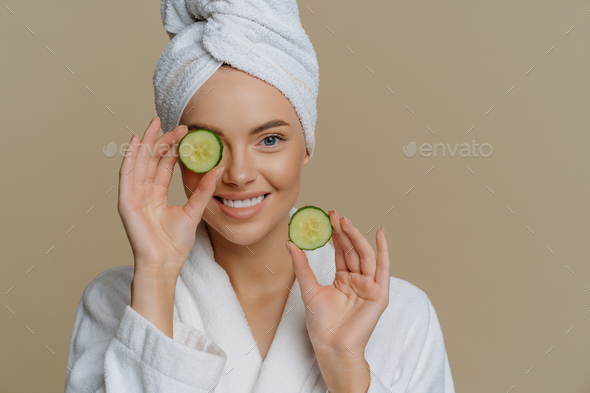 Positive refreshed beautiful woman holds slices of fresh cucumber makes mask for face skin
