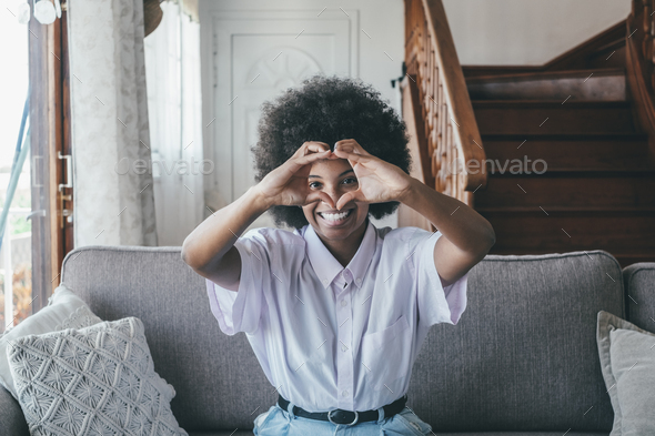 Happy african american woman sitting in living room connecting fingers of both hands making heart