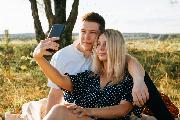 Happy young couple makes selfie on cell phone during romantic date outdoors, picnic in field at