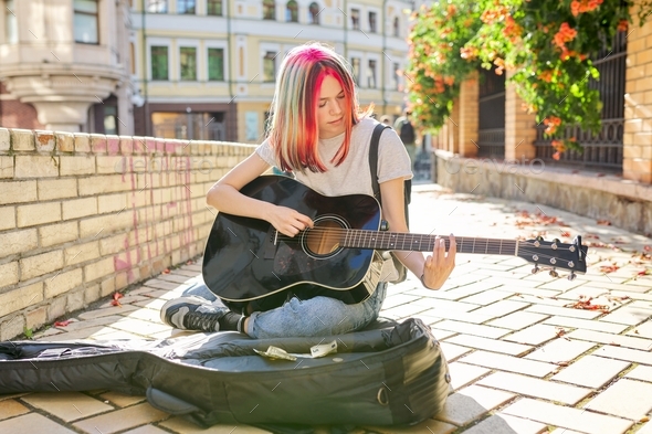 Creative female playing acoustic guitar sitting on sidewalk with guitar case and cash