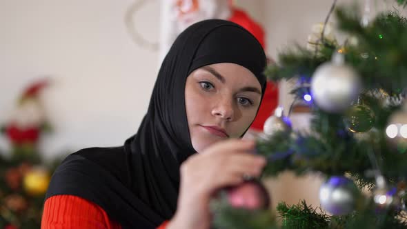 Closeup Young Caucasian Woman in Hijab Hanging Christmas Decoration Toys on New Year Tree alt