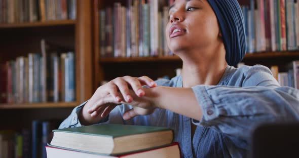 Asian female student wearing a blue hijab sitting and leaning on a pile of books and thinking alt