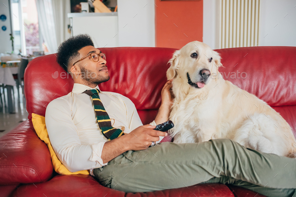 Young beautiful man indoor at home relaxing with his dog surfing channel using remote controller